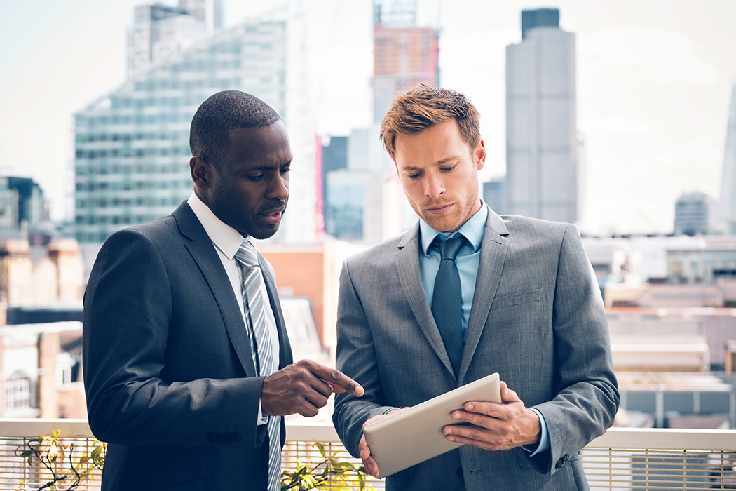 Two men in business suits standing in front of a city skyline looking at an iPad